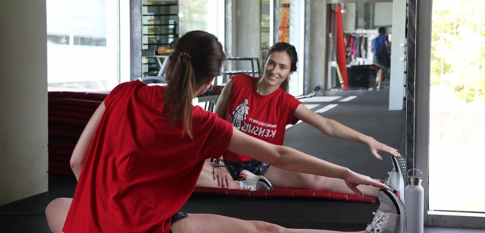 A woman in a red Crossjoin t-shirt sits on a gym mat stretching her leg while looking at her smiling reflection in a large wall mirror.