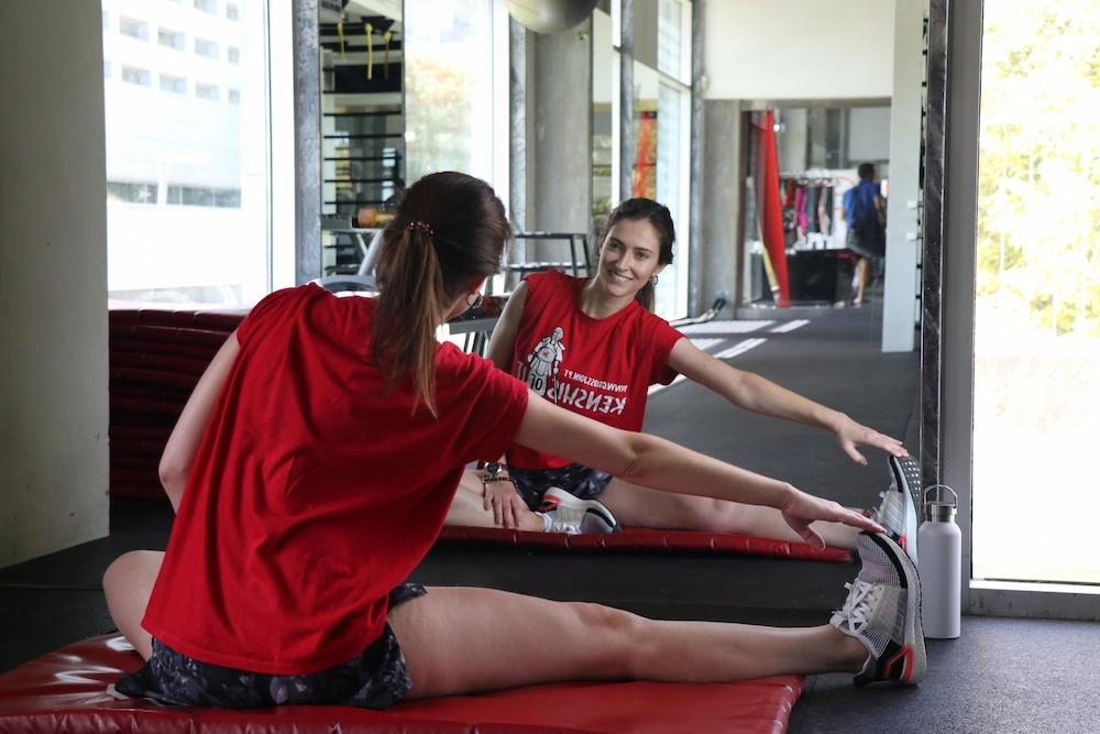 A woman in a red Crossjoin t-shirt sits on a gym mat stretching her leg while looking at her smiling reflection in a large wall mirror.